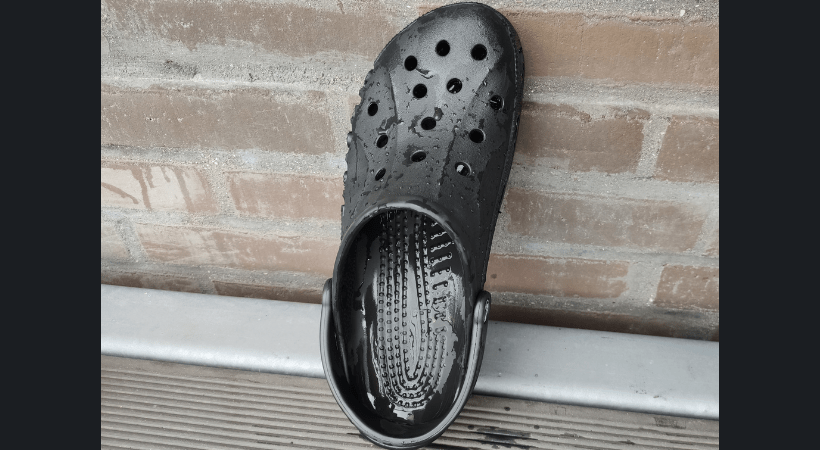 A black crocs clog is positioned vertically by the wall and left to air dry outside 